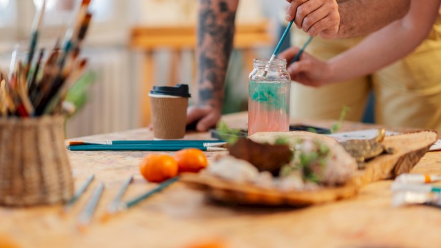 A messy art-and-snack table with brushes, a jar of water, a coffee cup, and a hand painting something while food sits nearby.