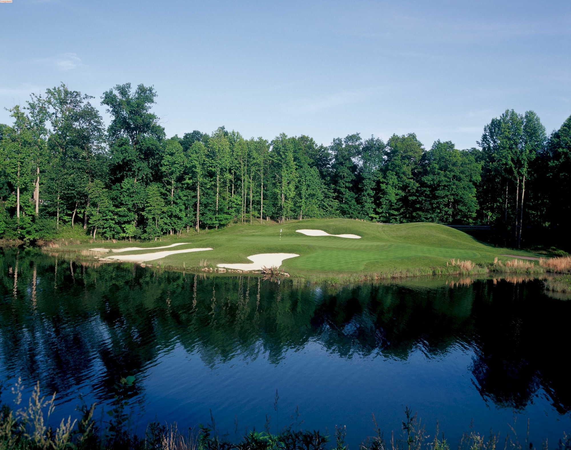 A serene golf course with green fairways and sand hazards is reflected in a calm lake, surrounded by a forest with clear blue skies above.