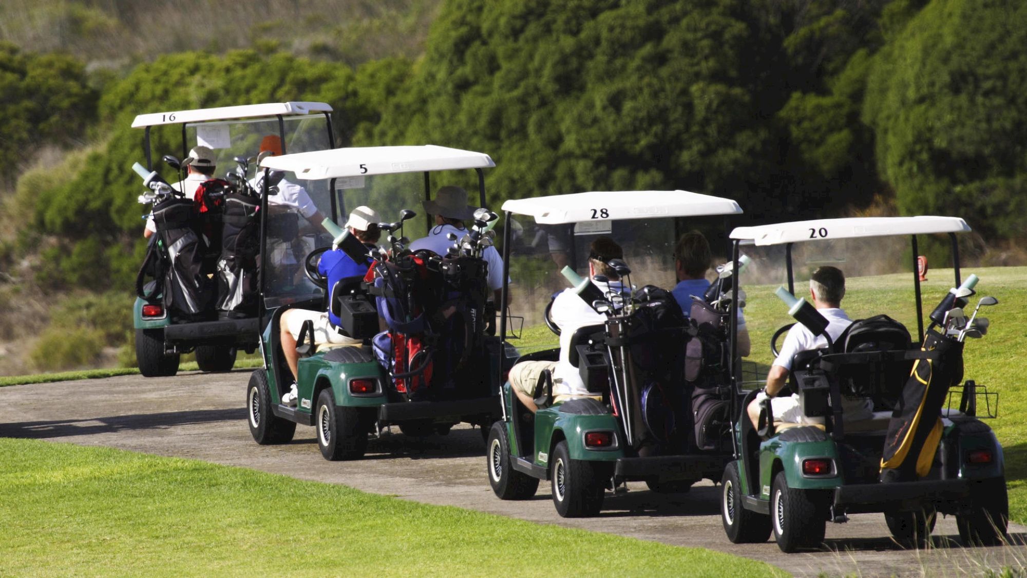People riding in golf carts on a golf course, with golf bags visible at the back of the carts and lush greenery in the background.