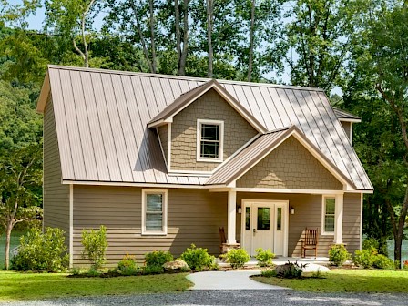 A cozy beige two-story house with a metal roof, front porch, surrounded by green trees and a lakeside yard, under a bright, sunny sky.