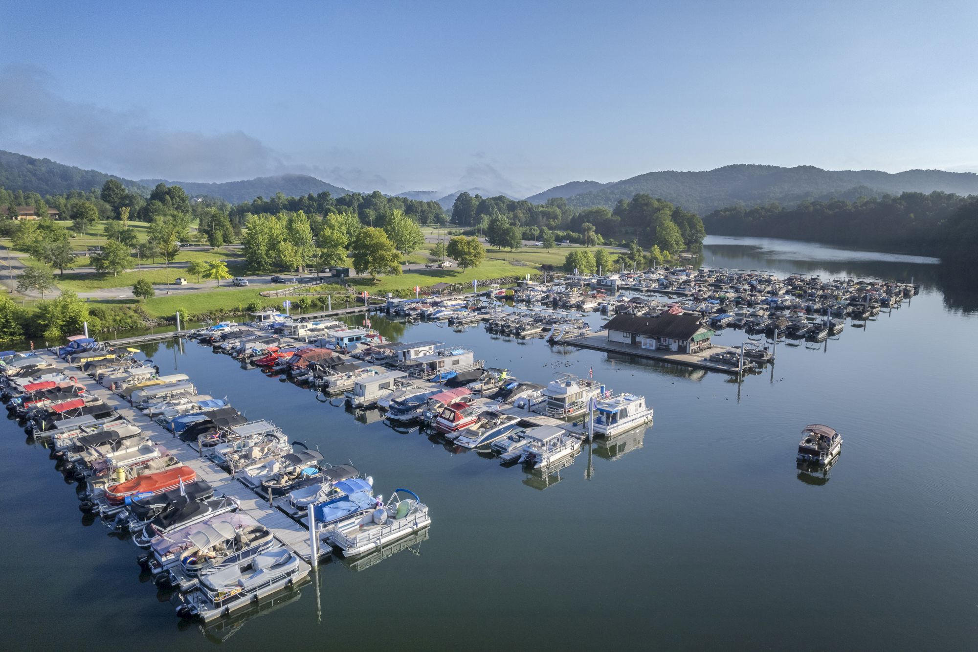 An aerial view of a marina with numerous boats docked in a calm, scenic body of water surrounded by green trees and distant hills.