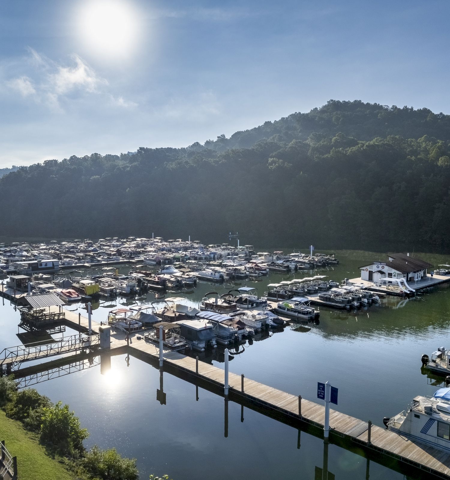 A marina filled with boats sits on a calm body of water surrounded by green hills under a bright sun, ending the sentence.