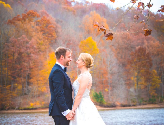 A newlywed couple stands by a lake with autumn-colored trees in the background, holding hands and smiling at each other.