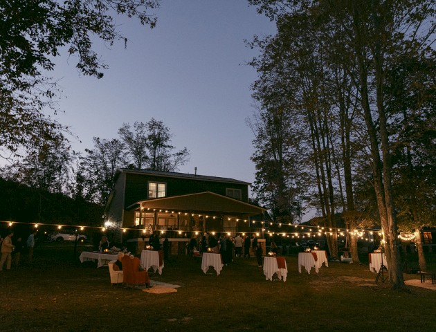An outdoor evening gathering with string lights, tables, and chairs in front of a house surrounded by trees.