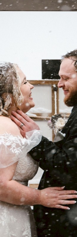 A joyful couple shares a romantic, snowy wedding moment, gazing at each other with happiness amid falling snow.