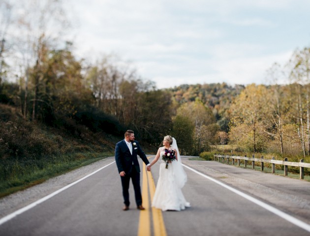 A bride and groom walk hand in hand on an open, deserted road surrounded by trees, celebrating their wedding day in a scenic outdoor setting.