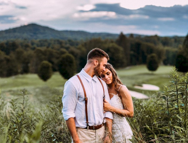 A couple embraces in a green field with mountains and cloudy sky in the background, conveying love, intimacy, and serenity.