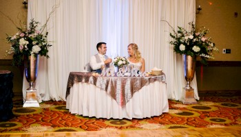 A couple sits at a decorated sweetheart table with flowers, white drapes, and tall floral arrangements flanking the scene, indoors at a wedding reception.