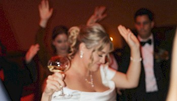 A bride in a white dress dances with a glass in hand, smiling and waving, surrounded by cheering guests.