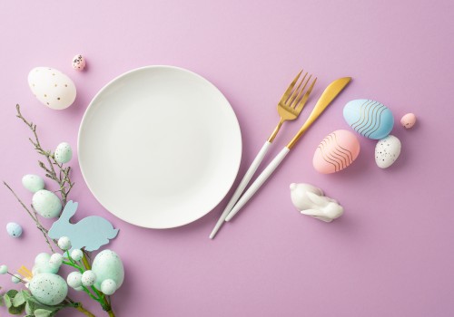 A white plate with a fork and knife on a pink background, surrounded by pastel Easter eggs, a small bunny tail, and spring greenery.