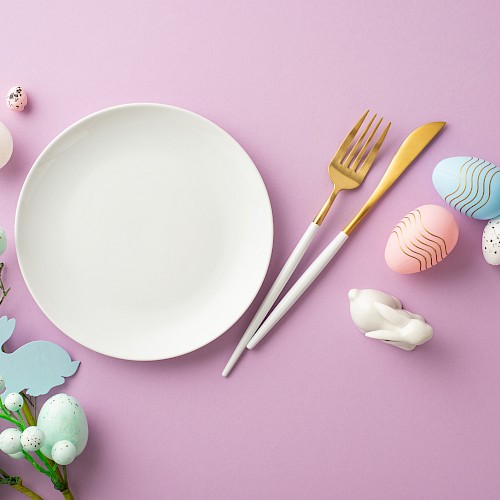 A white plate with a fork and knife on a pink background, surrounded by pastel Easter eggs, a small bunny tail, and spring greenery.
