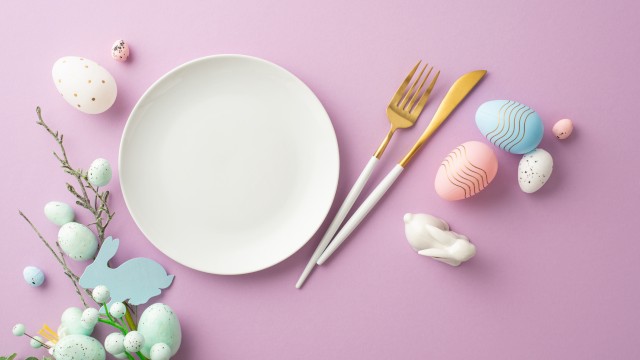 A white plate with a fork and knife on a pink background, surrounded by pastel Easter eggs, a small bunny tail, and spring greenery.