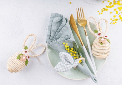 A pastel place setting with a mint plate, gray napkin, gold cutlery, a bow-tied napkin, and decorative woven eggs, plus yellow flowers, on a light background.