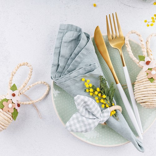 A pastel place setting with a mint plate, gray napkin, gold cutlery, a bow-tied napkin, and decorative woven eggs, plus yellow flowers, on a light background.