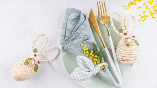 A pastel place setting with a mint plate, gray napkin, gold cutlery, a bow-tied napkin, and decorative woven eggs, plus yellow flowers, on a light background.