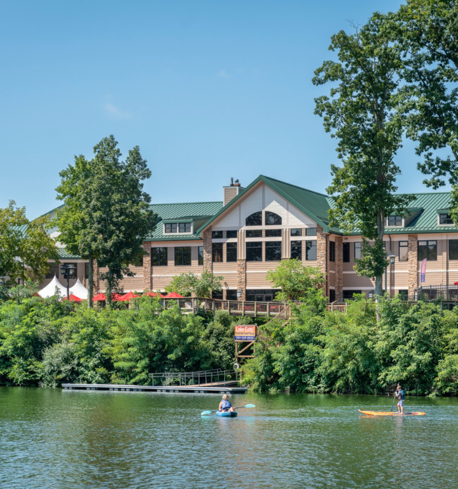 A large building by a river with people kayaking in the water, surrounded by lush greenery and trees under a clear blue sky.