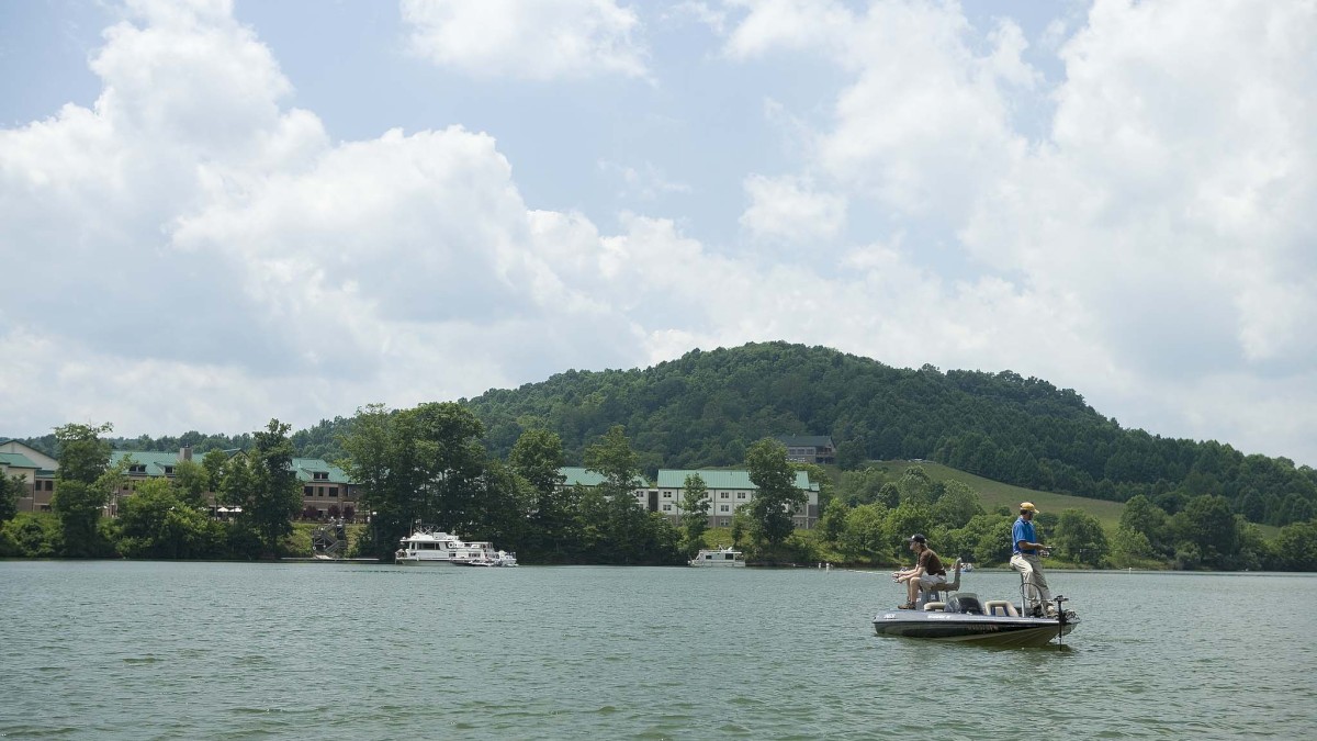 Two people are fishing on a small boat on a lake, with a hilly, forested area and buildings in the background under a partly cloudy sky.