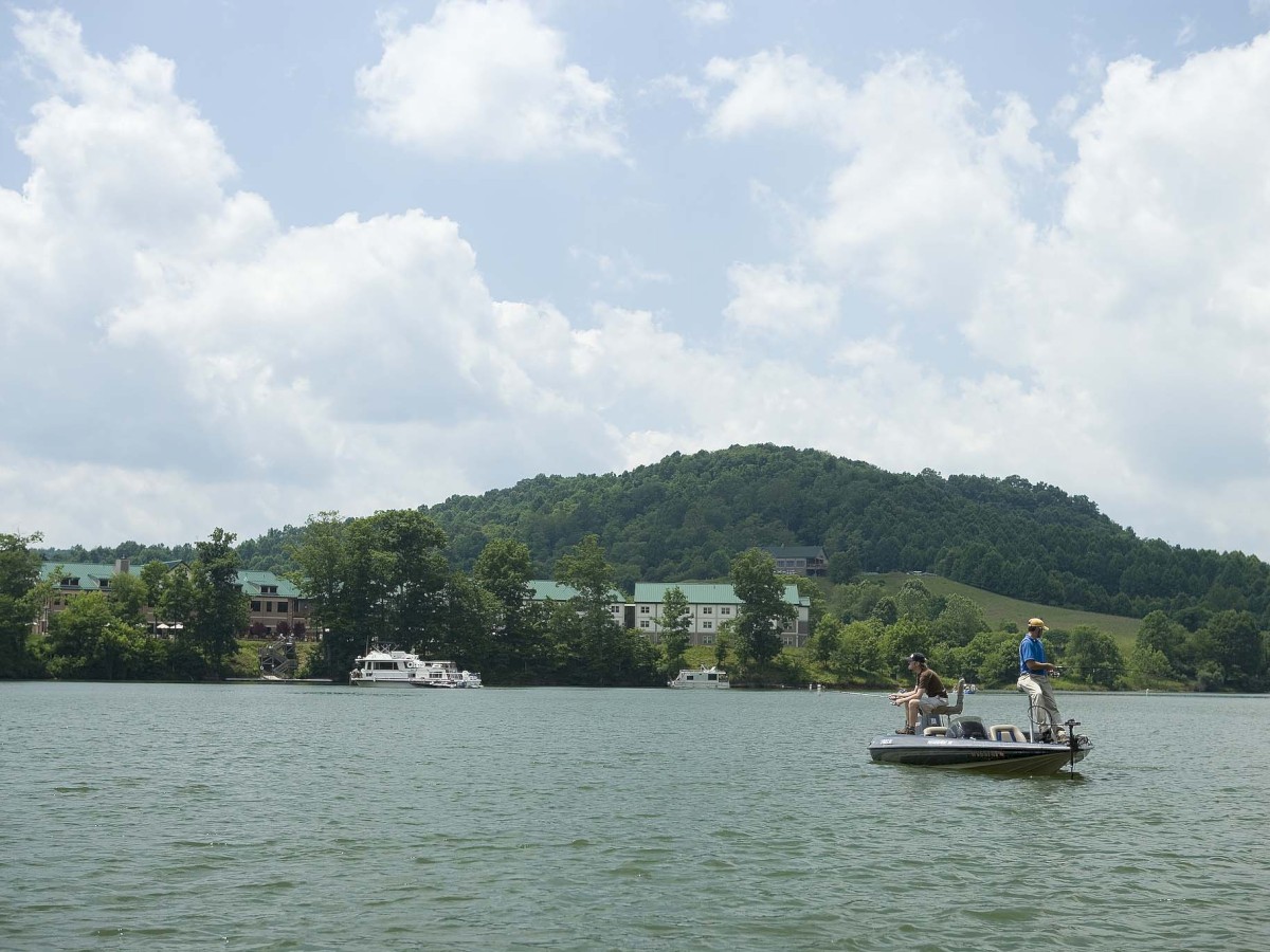 Two people are fishing on a small boat on a lake, with a hilly, forested area and buildings in the background under a partly cloudy sky.