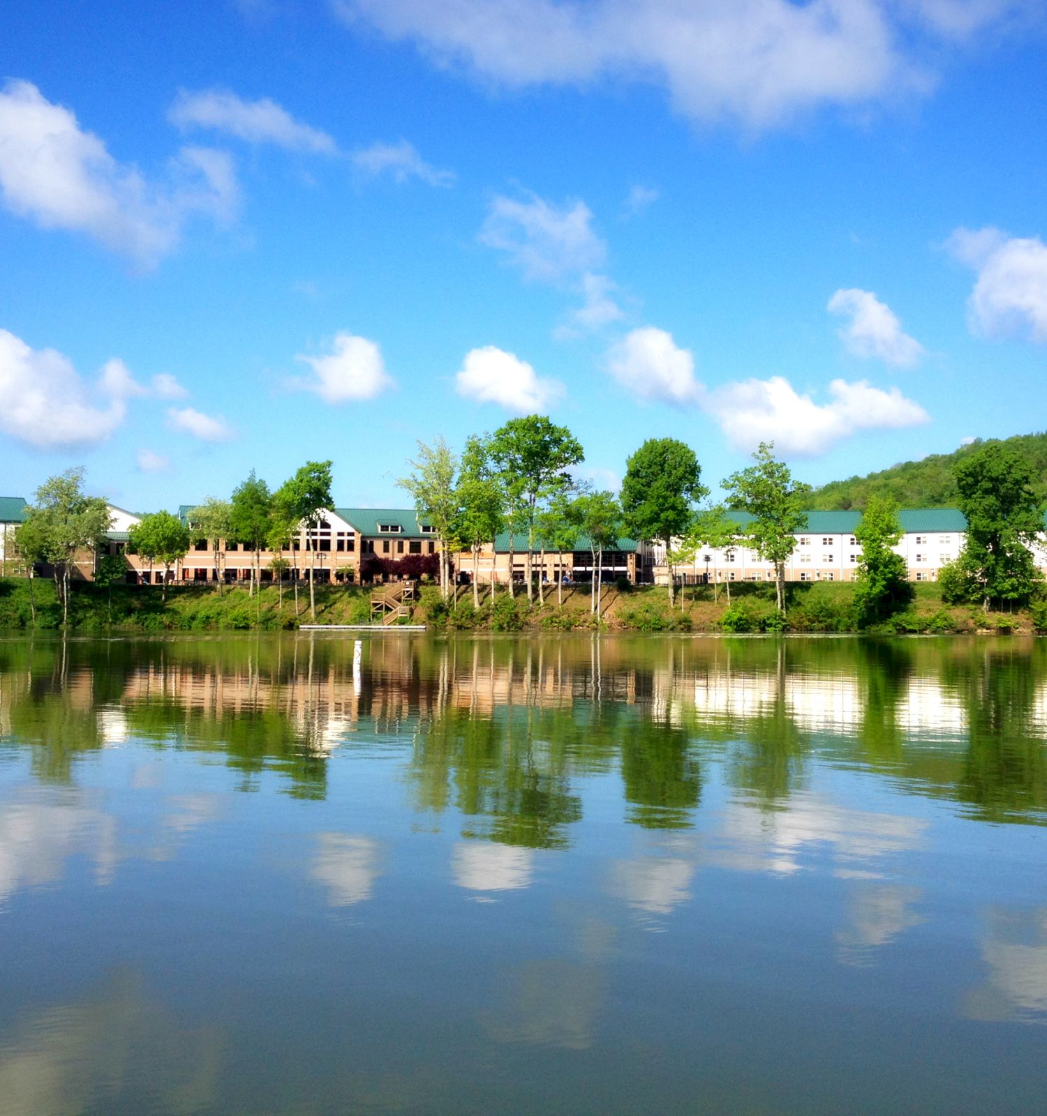 A serene lakeside view with buildings and trees reflected in the calm water under a bright blue sky with scattered clouds.