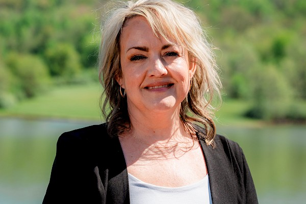 A woman with blonde hair stands outdoors near a lake, smiling, with green hills and a clear sky in the background.