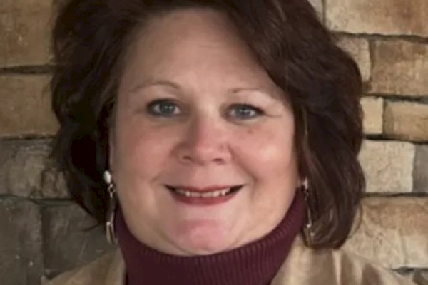 A smiling woman with short dark hair wearing a maroon turtleneck and beige jacket, posed in front of a stone wall.