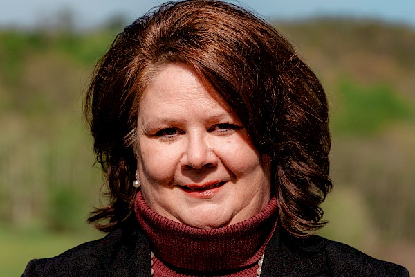 A woman with dark red hair on a sunny day, wearing a black blazer, maroon turtleneck, and a pearl necklace outdoors.