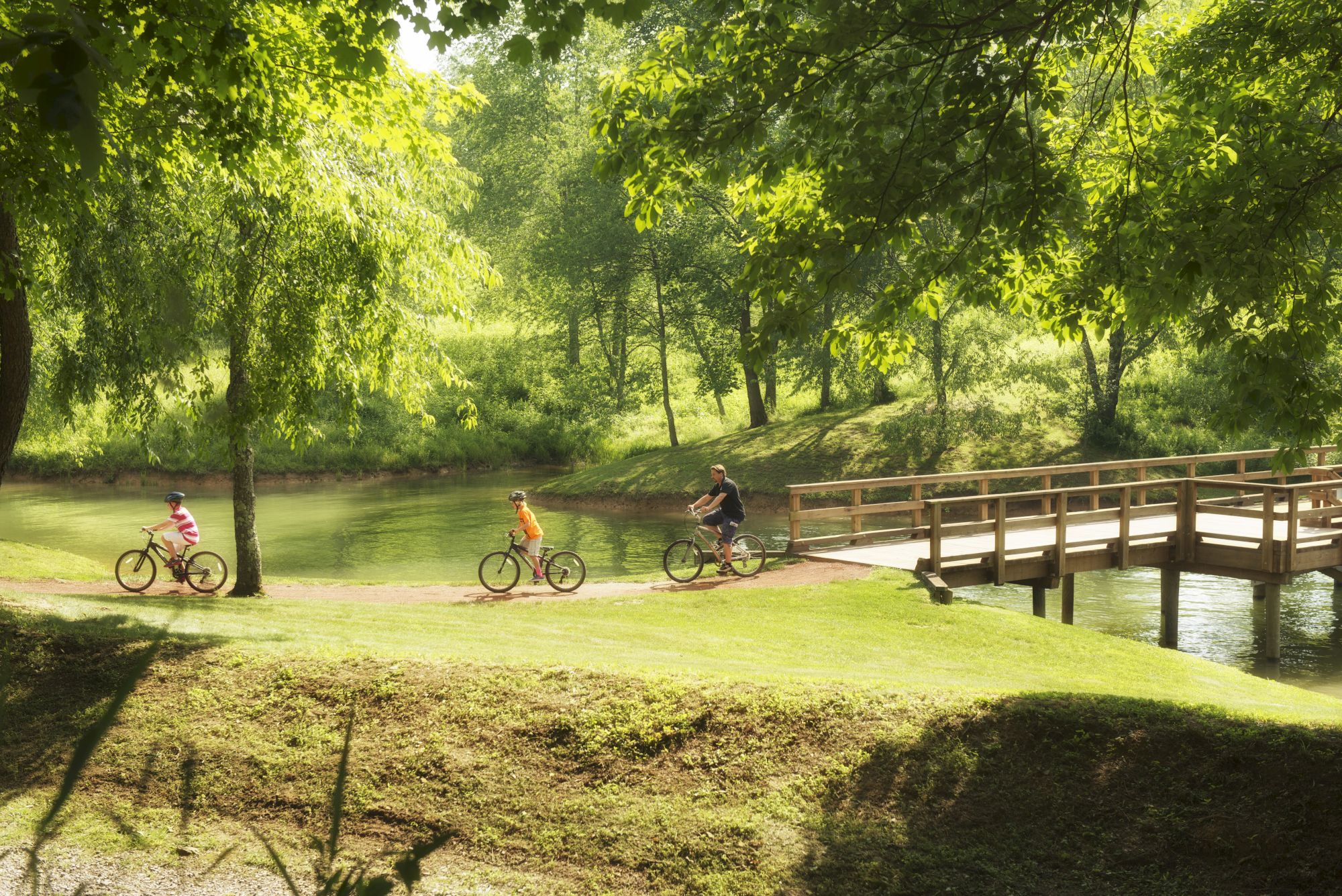 Three people are riding bicycles across a wooden bridge in a lush, green park with trees and a pond.