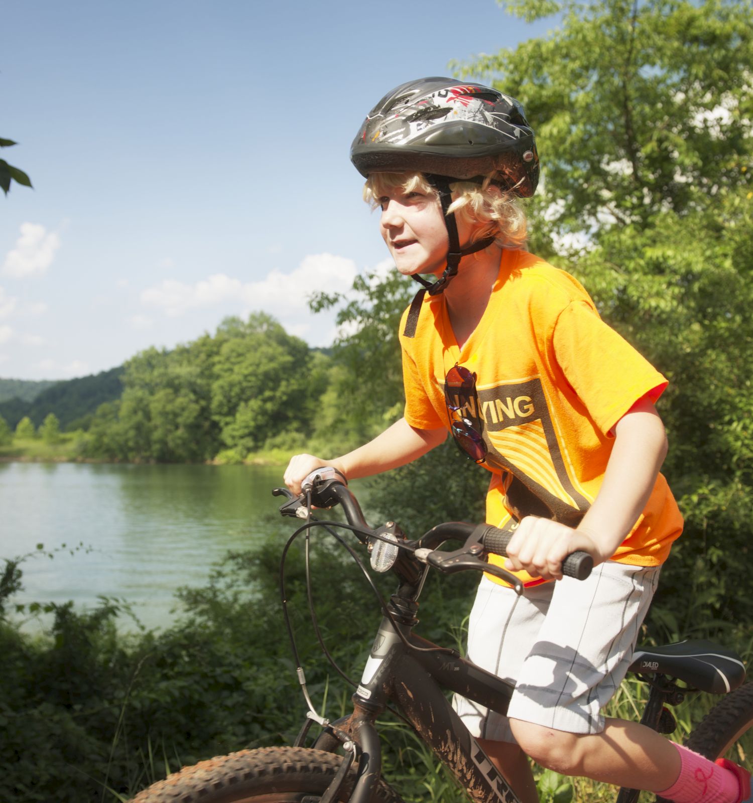 A child wearing a helmet and orange shirt rides a bicycle near a lake, surrounded by lush greenery and hills under a clear sky.