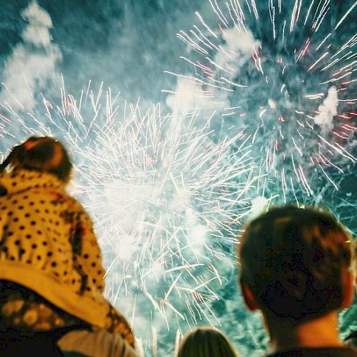 A crowd watches a fireworks display lighting up the night sky with bright bursts and trails, creating a festive, awe-filled atmosphere.