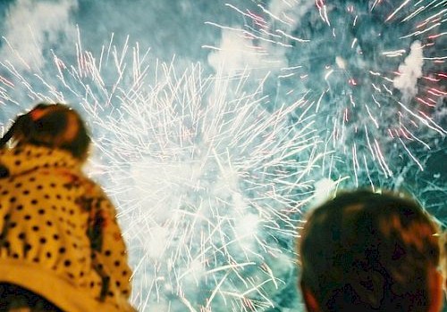Fireworks explode in the night sky while watching crowds, bright bursts lighting the silhouettes of two spectators.