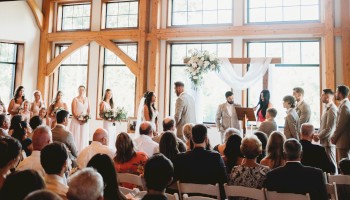 A wedding ceremony inside a bright, wooden-chateau style venue with arched windows, chandeliers, and guests seated facing the couple.