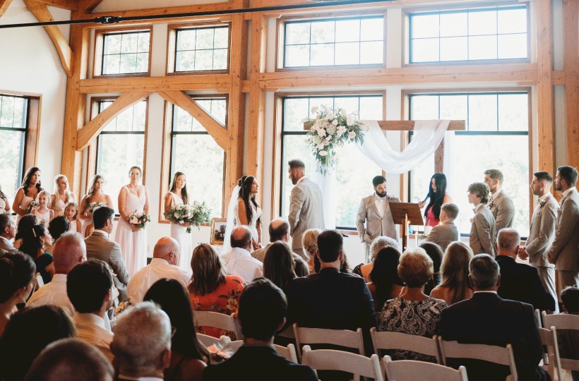A wedding ceremony inside a bright, wooden-chateau style venue with arched windows, chandeliers, and guests seated facing the couple.