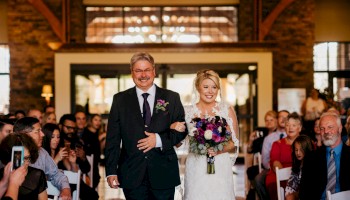 A bride and groom walk down the aisle arm-in-arm in a rustic church, wedding guests applaud as petals fall on the aisle.