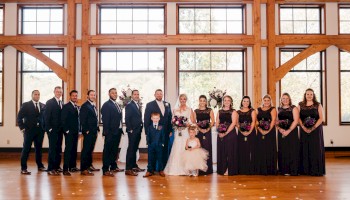 A wedding party stands in a bright hall: bride and groom center, attendants in black attire on either side, posing for a group photo.