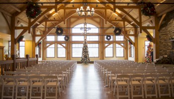 A rustic wedding chapel with wooden beams, rows of white chairs, a central Christmas tree, and festive wreaths under a high vaulted ceiling.