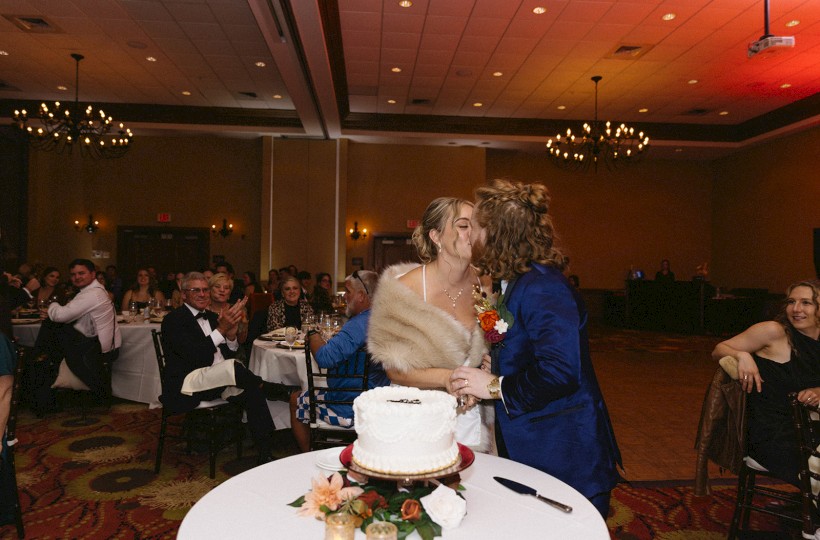 A couple kisses by a white wedding cake on a round table, guests seated in a banquet hall watching in the background.