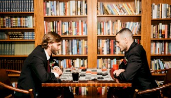 Two men in suits sit across a chessboard in a cozy library, focused on a game amid tall wooden shelves.