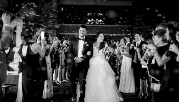 A bride in a voluminous white gown and groom in a suit walk arm-in-arm through a crowded, celebratory wedding reception with confetti and guests cheering, ending with a period.