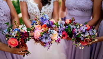 A bridal party with lavender dresses holds vibrant bouquets of mixed flowers, including pink, purple, and orange blooms, in a close, festive pose.