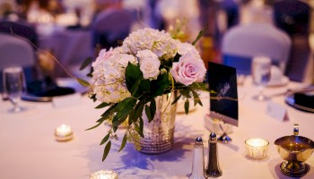 A floral centerpiece with pink and white roses in a metallic vase on a softly lit banquet table, surrounded by candles and place settings.