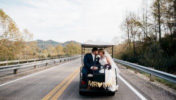 A couple in wedding attire sits in a small open vehicle on a rural road, 