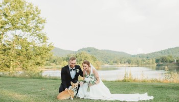 A bride and groom in wedding attire pose on a grassy lakeside with a dog, surrounded by trees and hills in the background.