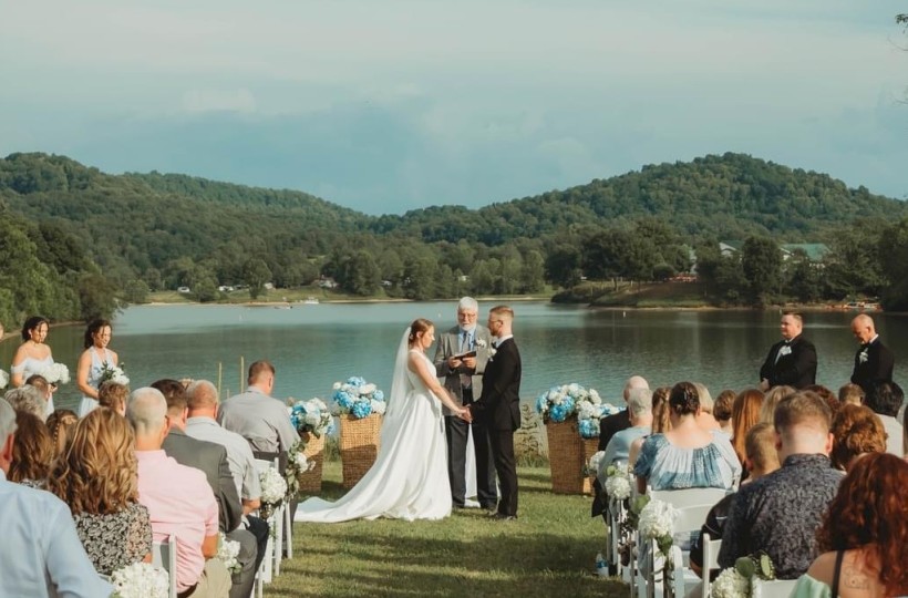 A wedding ceremony outdoors by a lake with hills, guests seated, couple at the altar under a clear sky.