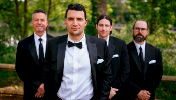 A stylishly dressed man in a black tux leads a group of three others in suits outdoors, all posing confidently for a formal photo.