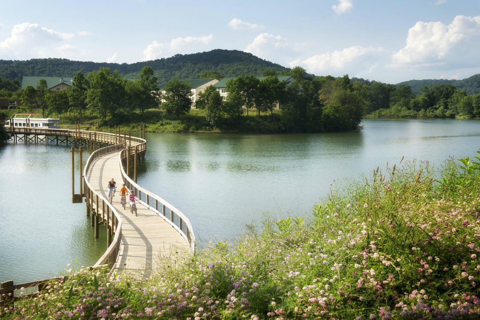 A wooden walkway curves over a lake, with people walking on it, surrounded by greenery and hills under a partly cloudy sky.