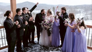 A wedding party gathers on a snowy balcony: brides in white and lavender, groomsmen in black, tossing toasts as bouquets sway.