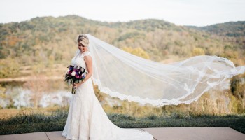A bride in a white gown holding a bouquet, standing outdoors with a long flowing veil billowing in the wind.