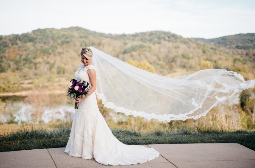 A bride in a white gown holding a bouquet, standing outdoors with a long flowing veil billowing in the wind.