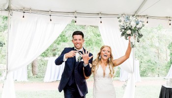 A newlywed couple walks under a white tent, bride in a white gown holding a bouquet, groom in a dark suit, both smiling and posing.