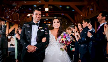 A bride and groom walk arm-in-arm down the aisle, smiling, as guests applaud in a warmly lit wedding venue.